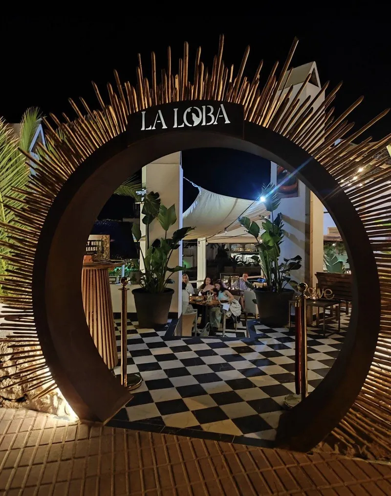 Arco de entrada de La Loba de noche, con corona de varas de madera iluminada y suelo de damero