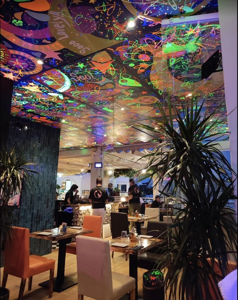 Sala principal con techo pintado de colores fluorescentes, palmeras y mesas de madera