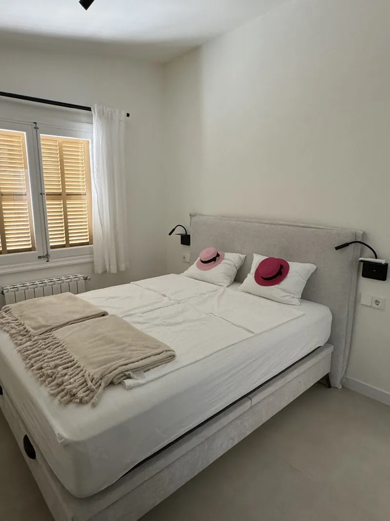 Dormitorio claro con cabecero tapizado, apliques de lectura y contraventanas