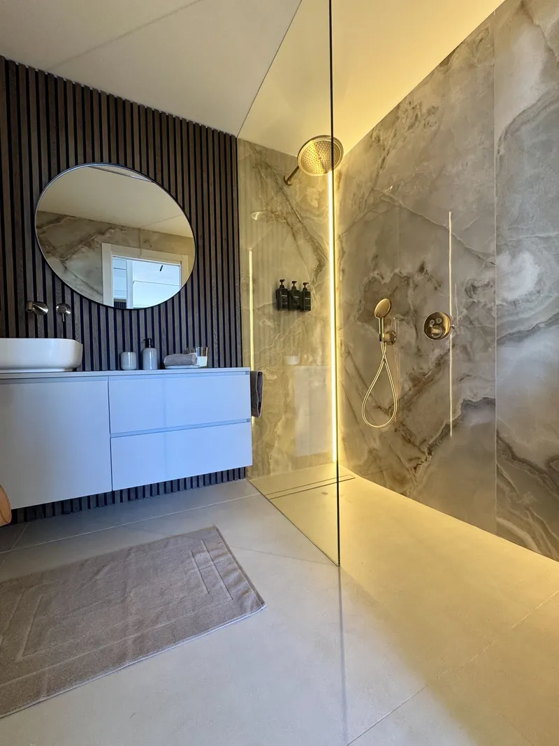 Baño con revestimiento de aspecto mármol, espejo redondo y ducha con luz lineal