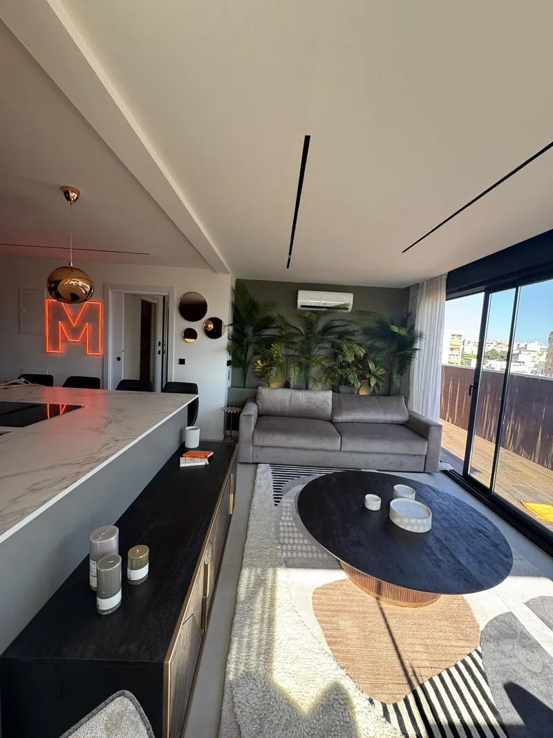 Zona de día con barra de cocina, letra luminosa, mesa ovalada y plantas al fondo