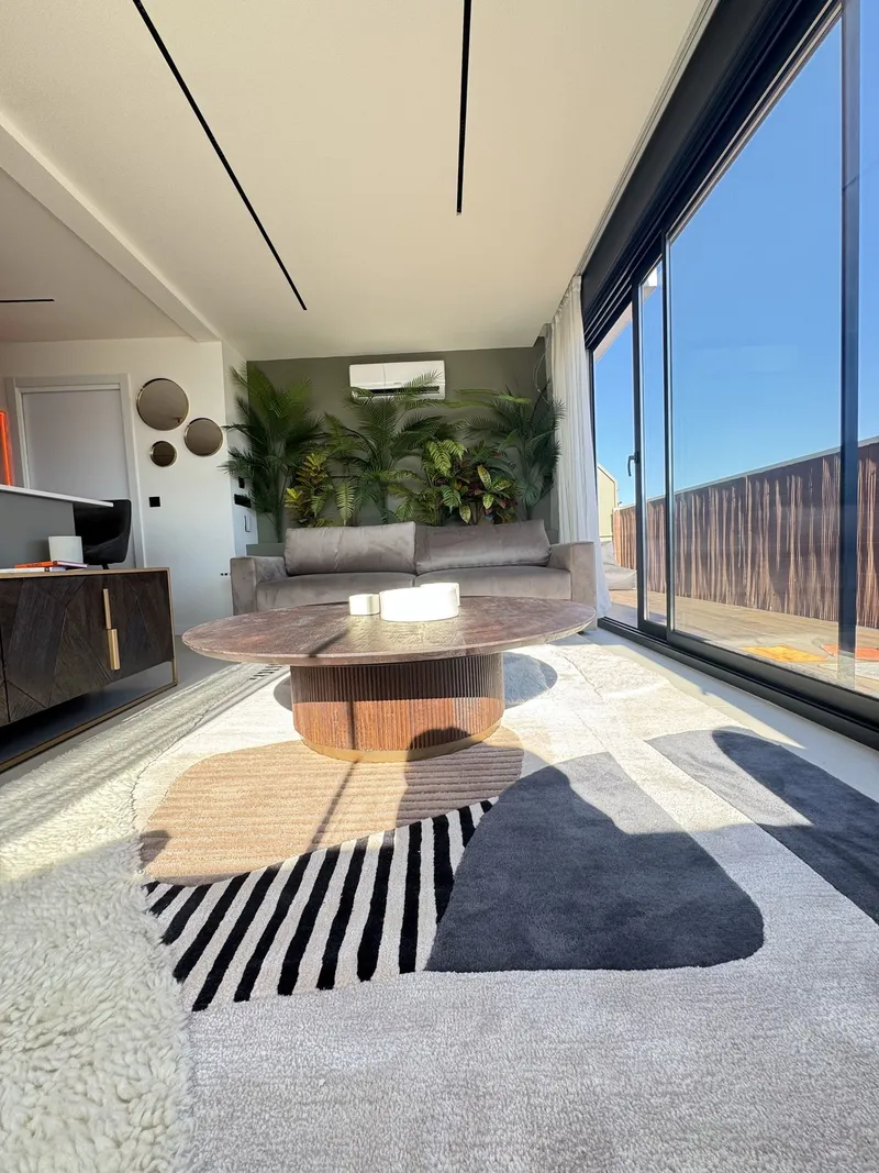 Salón con pared verde oliva, plantas, mesa de centro de madera y alfombra gráfica junto al ventanal