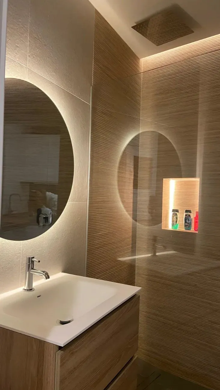 Baño con revestimiento de madera, espejos redondos retroiluminados y nicho iluminado