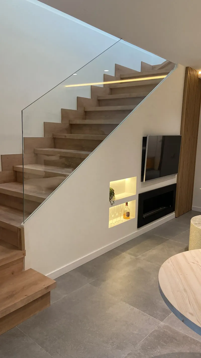Escalera de peldaños de roble con barandilla de cristal y tira LED, nicho iluminado y chimenea bajo la escalera