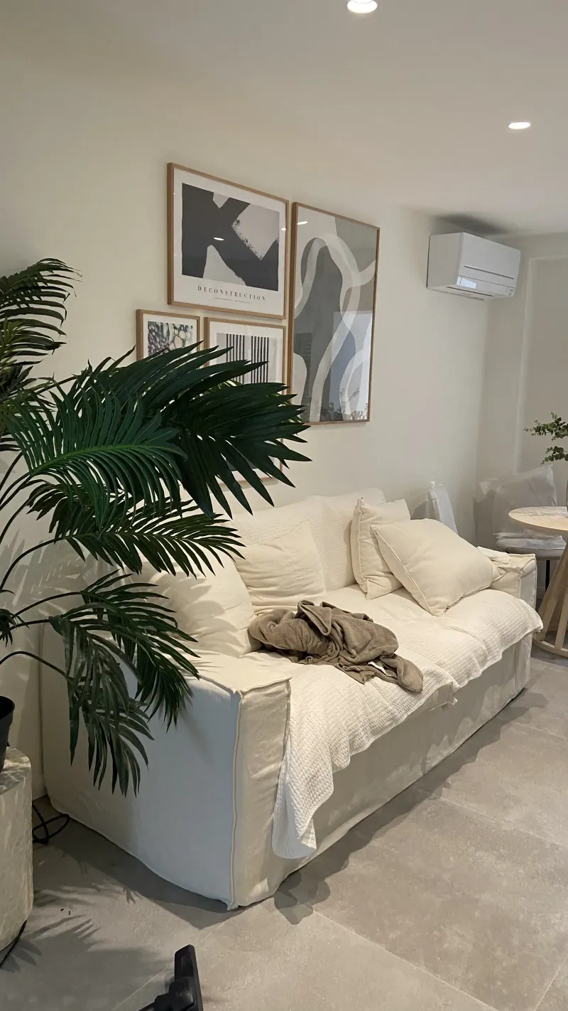 Salón en tonos arena y piedra con sofá de lino, palmera y láminas enmarcadas
