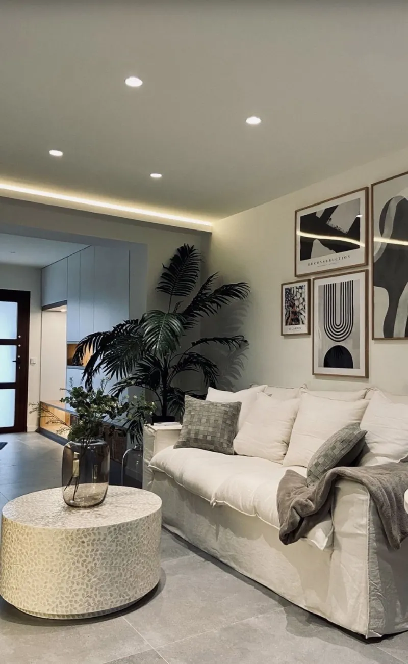Salón con sofá blanco, mesa auxiliar texturizada, palmera y láminas enmarcadas